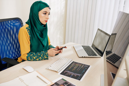 Serious pretty young muslim coder in traditional clothes reading e-mail on computer screen and...