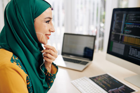 Cheerful smiling muslim young woman wearing earphones, programming on computer and talking to coworkerの写真素材