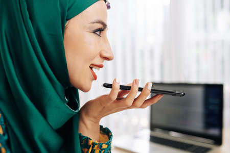 Cheerful beautiful muslim young businesswoman in hijab recording voice message for colleagueの写真素材