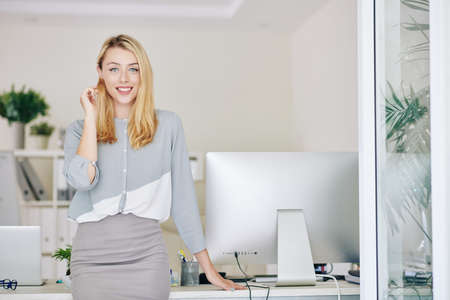 Beautiful smiling young female entrepreneur leaning on her office table and looking at cameraの写真素材