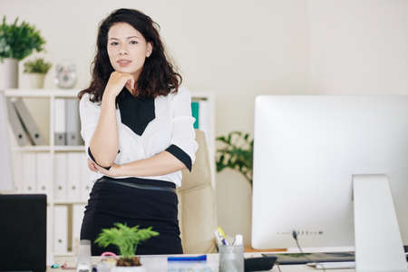 Portrait of pretty serious young businesswoman standing at office desk and looking at cameraの写真素材