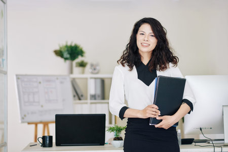 Beautiful young female entrepreneur with documents folder standing at her office desk