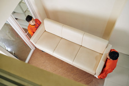 Movers carrying heavy beige leather sofa in new house, view from above