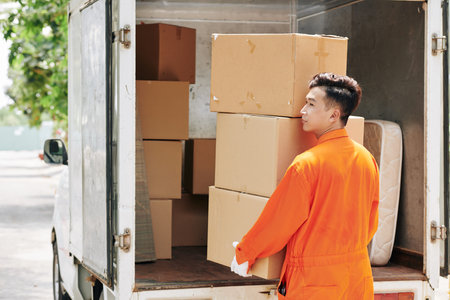 Asian young man carrying stack of cardboard boxes to truck trunk