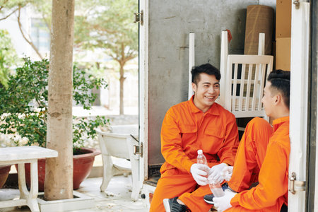 Cheerful young Vietnamese movers sitting in car trunk, drinking water and joking around after...