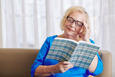 Smiling senior woman in glasses enjoying reading interesting novel when resting at home