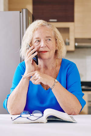 Aged woman sitting at kitchen table with opened book and talking on phoneの写真素材