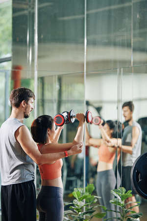 Fitness coach explaining young woman how to do lateral raise with dumbbells correctlyの写真素材