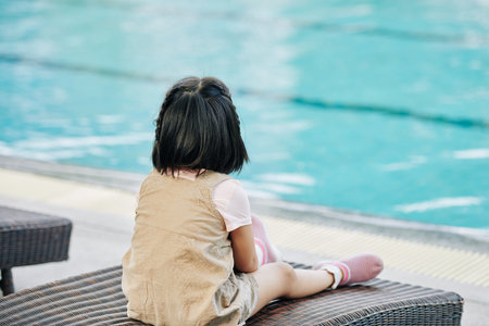 Pensive little girl sitting on chaise-lounge and looking at turquoise water in swimming poolの写真素材