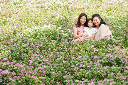 Cheerful mother, grandmother and granddaughter sitting among blooming flowers on meadow