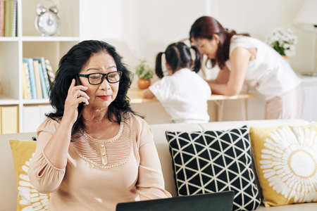 Senior Vietnamese woman in glasses talking on phone when reading business document on laptop screen when staying at home with her familyの写真素材