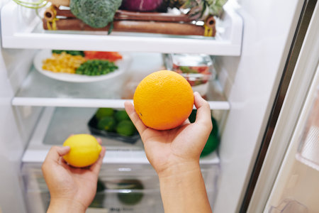 Woman taking citrus fruits: orange and lemon out of refrigerator when searching for snacks at home