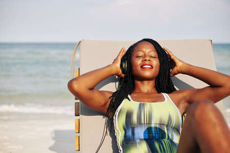Happy beautiful young Black woman relaxing in chaise-lounge and listening to music in headphonesの写真素材