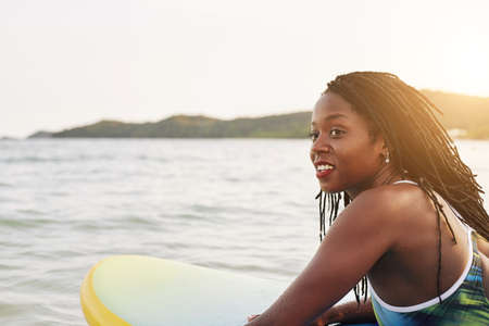 Beautiful smiling young Black woman sub surfing in ocean under rays of sunset sunの写真素材