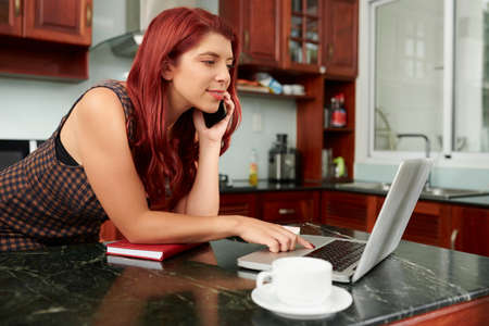 Pretty smiling young woman staying home during quarantine, making orders in online store and talking on phoneの写真素材