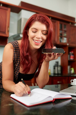Positive young woman recording voice message when taking notes in planner on kitchen counterの写真素材