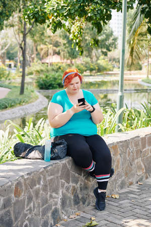 Young plus size woman resting on stone wall in park and checking messages and notifications in her smartphoneの写真素材