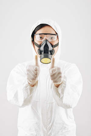 Portrait of serious Asian man in white hazmat suit and protective mask showing thumbs-up and looking at cameraの写真素材