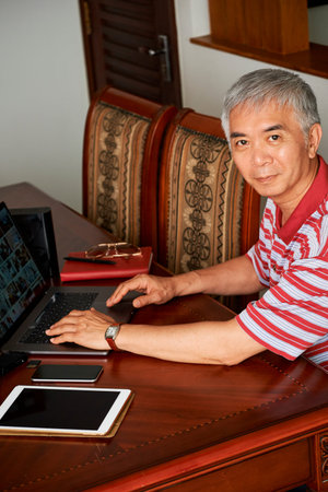 Elderly Chinese man working on laptop at home and editing photos he made on his new camera