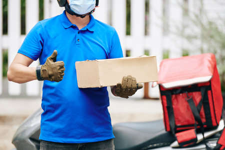 Delivery man with cardboard box standing at his scooter and showing thumbs-upの写真素材