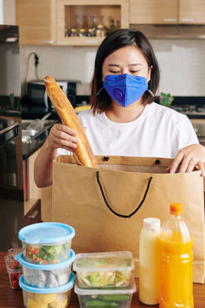 Young Asian woman in medical mask opening paper bag after receiving food delivery and taking out fresh bread, drinks, and vegetablesの写真素材