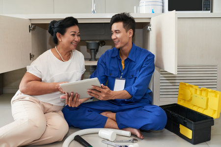 Smiling plumber showing senior woman tablet computer and explaining how he will repair kitchen drain...