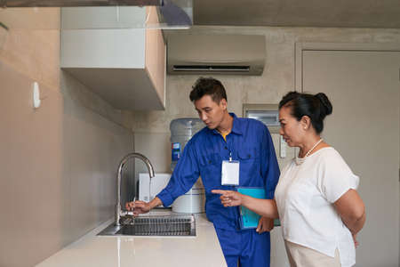 Senior Vietnamese woman showing leaking sink tap in kitchen to plumber in blue uniformの写真素材
