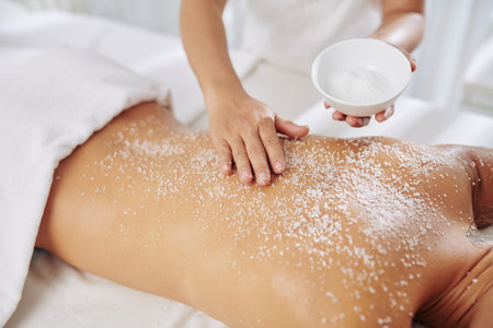 Beautician exfoliating back of young woman with salt scrub before massageの写真素材
