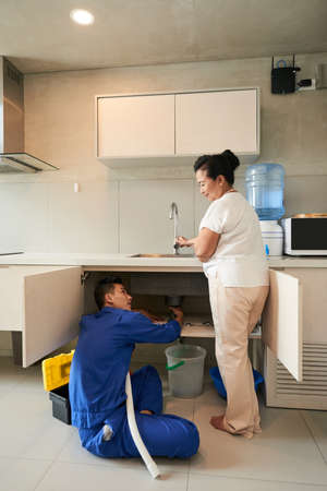 Senior woman turning on faucet when plumber checking leaking pipe of kitchen sinkの写真素材