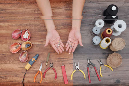 Hands of woman showing new earrings she made with tools, threads and beads around, view from aboveの写真素材