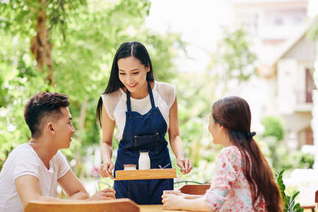 Pretty smiling young waiter bringing tray with organic non-dairy yogurt to couple sitting at cafe tableの写真素材