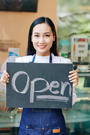Positive young Asian woman in denin apron standing in cafe and showing open signの写真素材