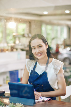 Portrait of young smiling Vietnamese cafe owner working on tablet computer and taking notes in plannerの写真素材
