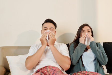 Young Vietnamese couple lying in bed and sneezing in handkerchiefの写真素材