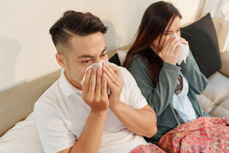 Young sick Asian couple sitting in bed under blanket and blowing noses in tissues at homeの写真素材