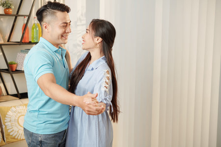 Beautiful young Asian man and woman looking at each other with love when dancing at homeの写真素材