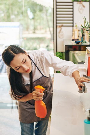 Pretty young coffeeshop owner sparying detergent on surfaces when disinfecting counter