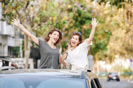Joyful happy pretty young women raising hands when riding in car trunkの写真素材