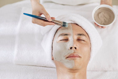 Cosmetologist applying clay mask on half of face of male clientの写真素材