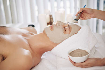 Hands of beautifcian applying mud mask on face of young man relaxing in beauty salonの写真素材