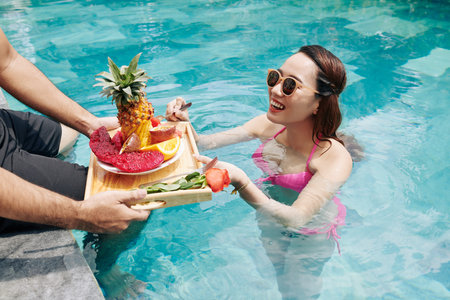 Happy pretty young woman in swimming pool taking piece of fruit from tray with breakfast that...