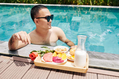 Happy young Asian woman standing in swimming pool and eating delicious breakfastの写真素材