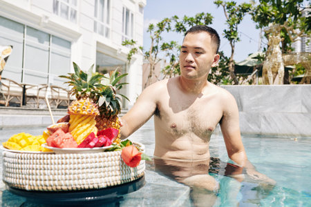 Handsome young Vietnamese man sitting on edge of swimming pool and eating tasty ripe fruits