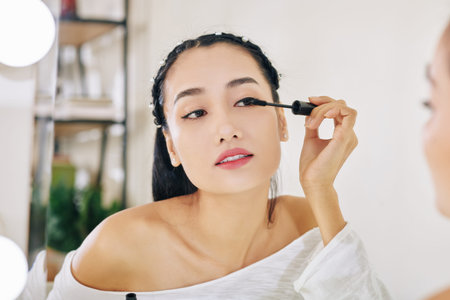 Pretty young Asian woman looking at mirror and applying second coat of black mascaraの写真素材