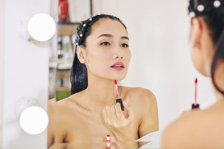 Beautiful young Asian woman looking at mirror and applying red lip glossの写真素材