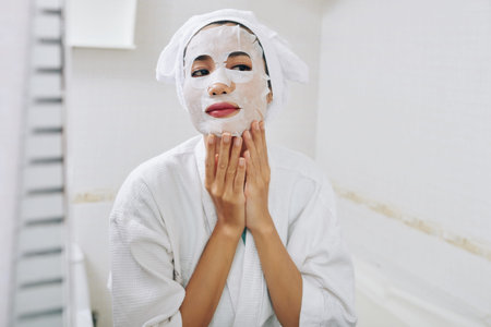 Beautiful young woman applying sheet mask on her face after taking shower