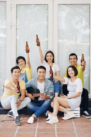 Hands young man playing guitar when his happy friends raising hands with bottles of beerの写真素材