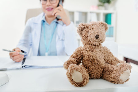 Teddy bear on desk of pediatrician talking on phone and filling documentsの写真素材