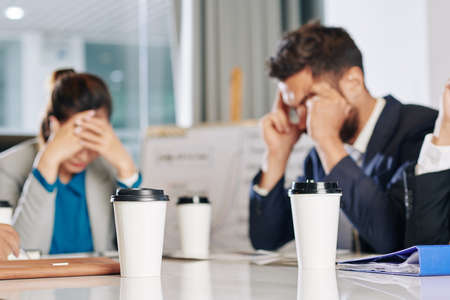 Cups of take out coffee on table in front of business people meeting to discuss economic consequences of covid-19 pandemicの写真素材