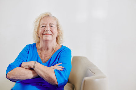 Portrait of smiling aged woman in blue dress crossing arms and looking at camera when sitting in chairの写真素材